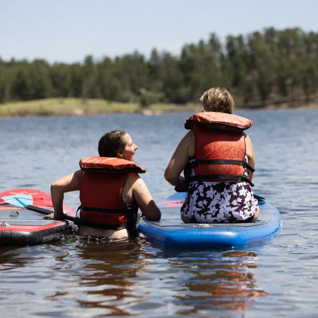Two individuals faced away from the camera look at each other in conversation. They are in a body of water and wearing life jackets. One is standing in the water and the other is sitting on a paddleboard.